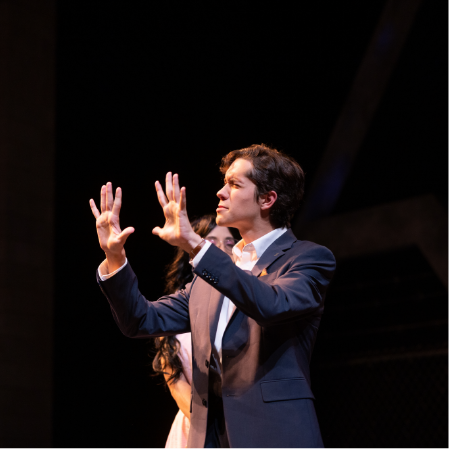 Two people on stage, one in the foreground gesturing with open hands. The face is obscured by a brown rectangle. Stage lighting and a black backdrop suggest a performance